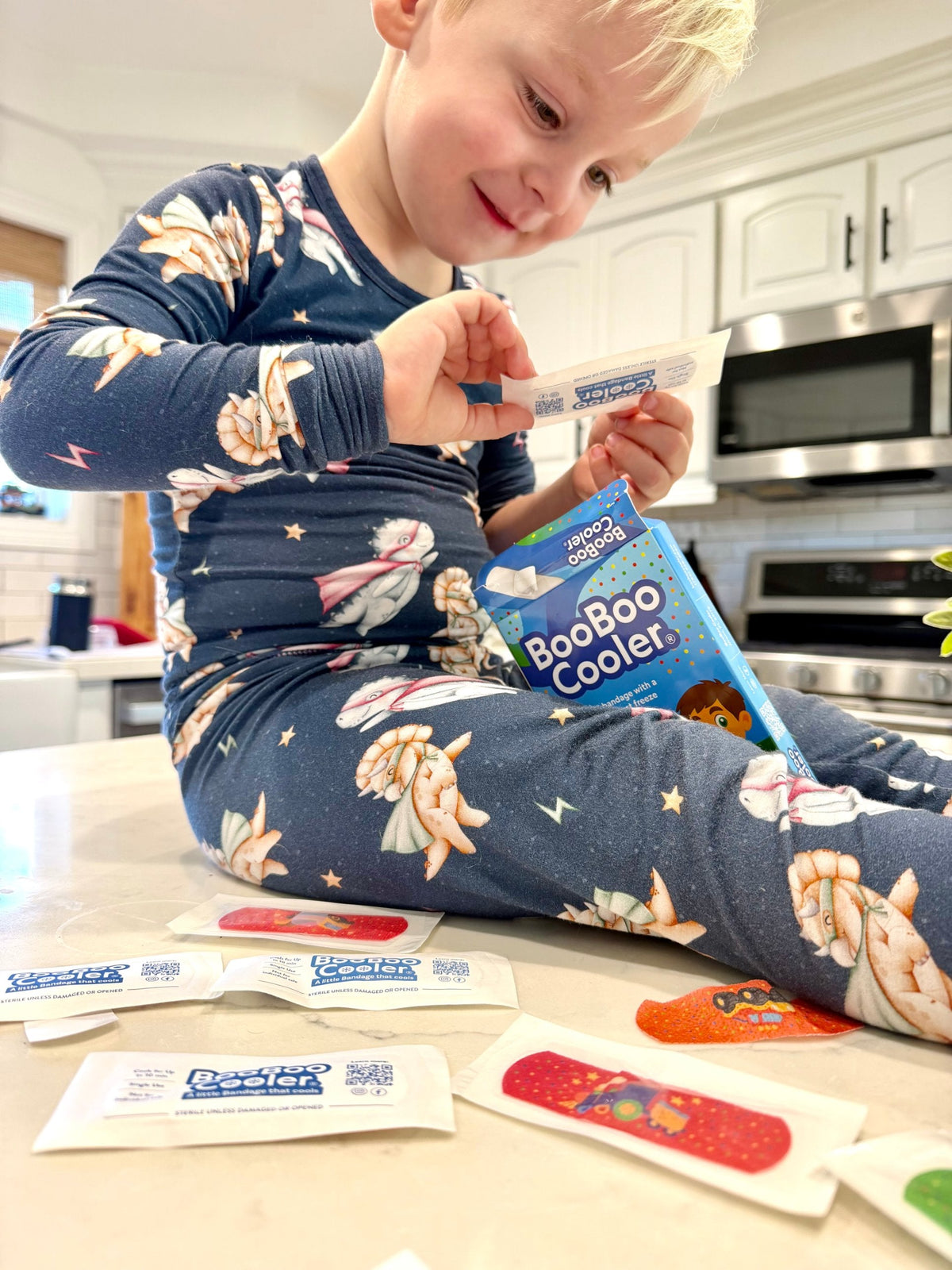 A toddler in dinosaur pajamas unwraps a BooBoo Cooler® bandage, with the blue BooBoo Cooler® box and individually sealed sterile bandages on the counter beside him.