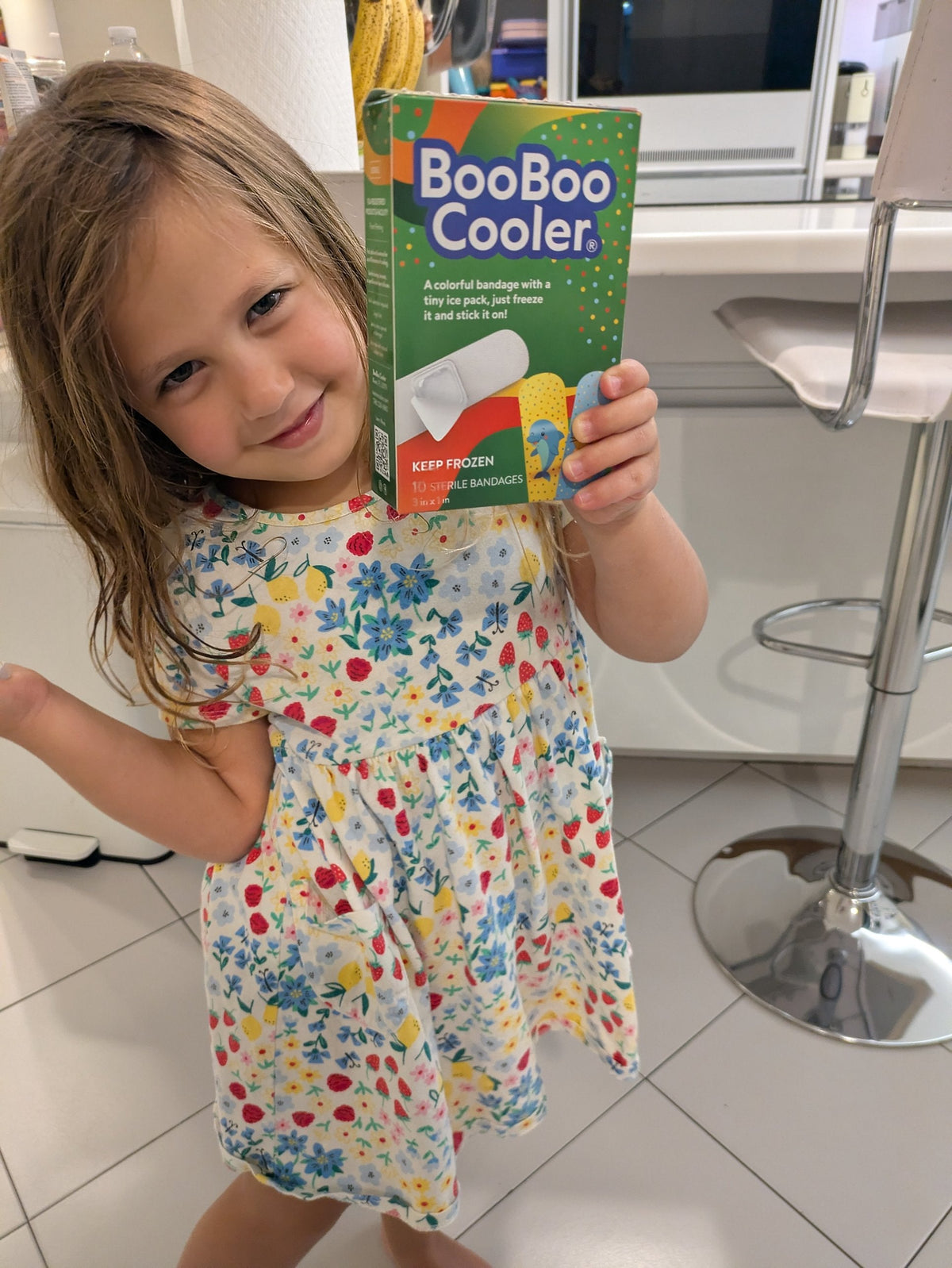 A smiling young girl in a floral dress holds up a green BooBoo Cooler® box like she just received a gift.