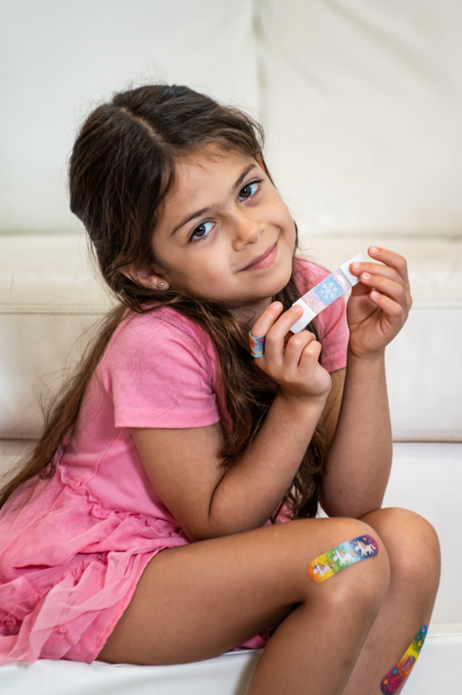 Young girl in a pink dress holding a bandage with colorful designs on a white couch.