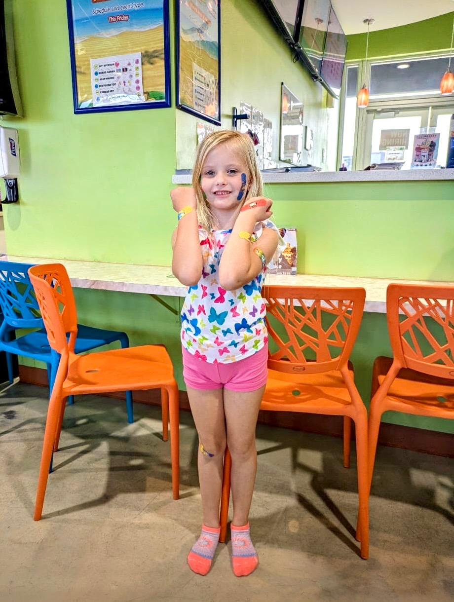 A young girl stands in a vibrant children’s play gym, adorned with orange chairs and green walls. She proudly displays her easily applied cooling bandages, a testament to the rough day of play she’s endured.