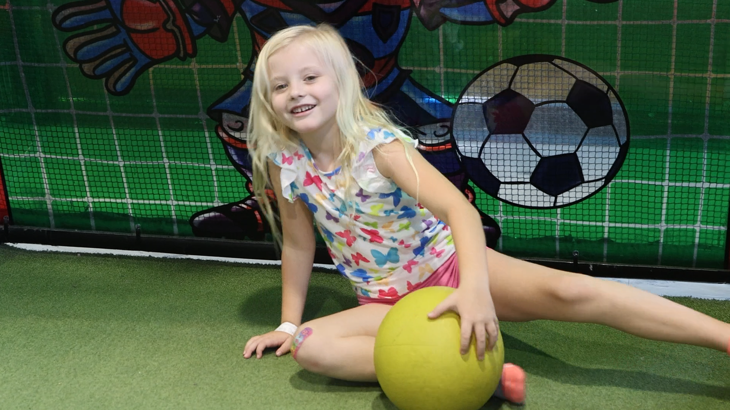 Young girl with a yellow ball in an indoor play area with sports-themed decorations while proudly displaying her cooling bandage on the knee.